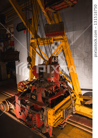Industrial machine marked 43 and 45 under a yellow crane with chains, set against a wall with technical drawings and warning signs about suspended loads. Industrial machine marked 43 and 45 under a yellow crane with chains, set against a wall with technical drawings and warning signs about suspended loads. 131518750