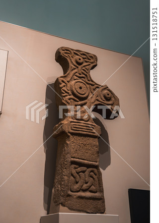 A detailed stone cross featuring Celtic style spirals, knots, and motifs, mounted on a pedestal in a museum or gallery with focused lighting. A detailed stone cross featuring Celtic style spirals, knots, and motifs, mounted on a pedestal in a museum or gallery with focused lighting. 131518751