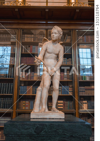 A marble statue of a winged figure holding a bow on a green pedestal, set in a well lit library with bookshelves and glass fronted cabinets. 131518764