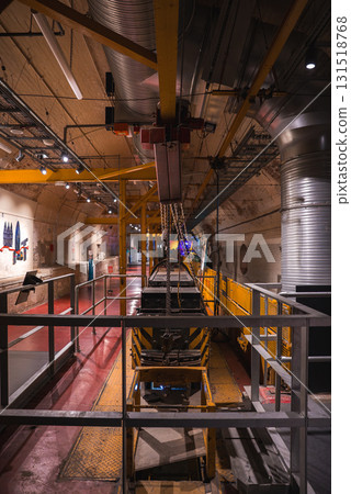 Tunnel like industrial interior featuring yellow metal beams, chains, a conveyor system, curved walls with pipes, and informational displays in London. Tunnel like industrial interior featuring yellow metal beams, chains, a conveyor system, curved walls with pipes, and informational displays in London. 131518768