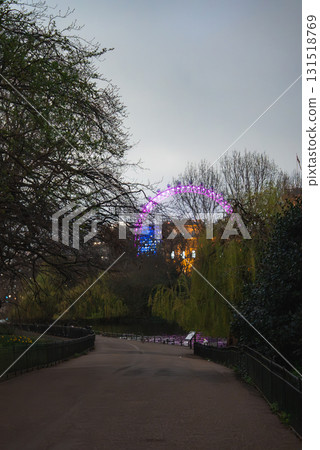 A tranquil park pathway in London bordered by trees and greenery, with the London Eye glowing in purple and blue against a dim evening sky. A tranquil park pathway in London bordered by trees and greenery, with the London Eye glowing in purple and blue against a dim evening sky. 131518769