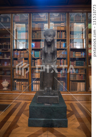A dark stone statue of the goddess Sekhmet with a lioness head, seated on a throne, displayed in a library like room with wooden bookshelves and polished floors. 131518773