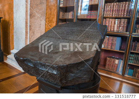 The Rosetta Stone on a pedestal in a museum room with wooden floors, marble columns, and bookshelves filled with historical books. The Rosetta Stone on a pedestal in a museum room with wooden floors, marble columns, and bookshelves filled with historical books. 131518777