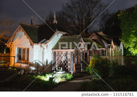 A cottage style building with pointed gables and decorative woodwork, surrounded by a garden with lattice fencing, set against bare trees at night. A cottage style building with pointed gables and decorative woodwork, surrounded by a garden with lattice fencing, set against bare trees at night. 131518781