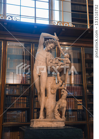 A marble statue of a male figure holding a fruit basket, with a child and dog at his feet, displayed indoors among tall wooden bookshelves and large windows. A marble statue of a male figure holding a fruit basket, with a child and dog at his feet, displayed indoors among tall wooden bookshelves and large windows. 131518782