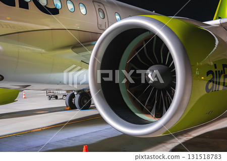 Modern airplane engine with green and white colors, airBaltic branding, fan blades, safety cones, and ground equipment on a lit tarmac at night. 131518783