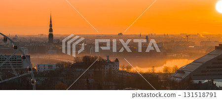 A warm sunrise illuminates Riga's old town with St. Peter's Church spire, historic rooftops, the Daugava River, and the National Library of Latvia. 131518791