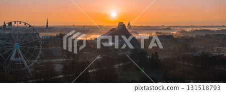 A warm sunrise illuminates Riga, Latvia, featuring the National Library, St. Peter's Church spire, a Ferris wheel, and the misty Daugava River. 131518793