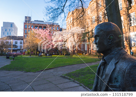 A serene park in Manchester featuring a bronze statue, red brick buildings, cherry blossom trees, and a mix of modern and traditional architecture. 131518799