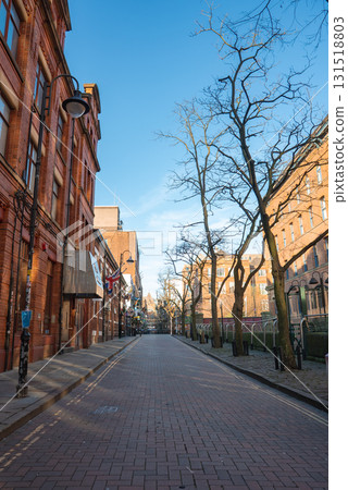 A red brick paved street in Manchester, England, lined with red brick buildings, bare trees, and a mix of modern and historic architecture under a clear sky. 131518803