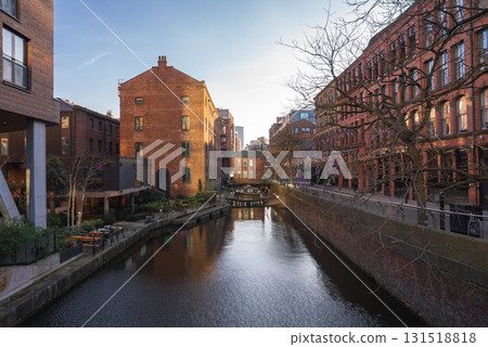 A tranquil canal in Manchester, England, bordered by historic red brick buildings, tree lined pathways, and outdoor seating under warm sunset lighting. 131518818