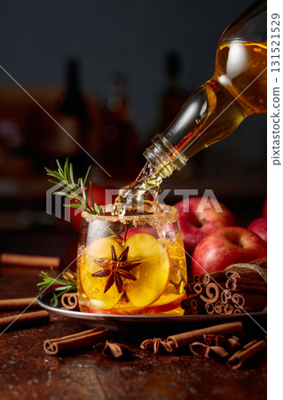 Apple cider is poured from a bottle into a glass with apple slices, anise, and a rosemary branch. Apple cider is poured from a bottle into a glass with apple slices, anise, and a rosemary branch. 131521529
