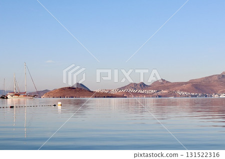 Sailboats and yachts rest on calm Mediterranean waters with dry coastal hills and scattered white homes in the background under a clear blue sky in Bodrum. 131522316