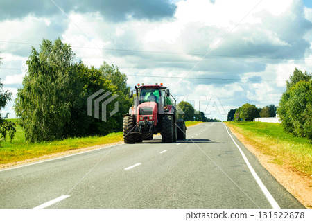 A red modern tractor with a semi-trailer and a barrel for watering plants on a field against the background of a village road and sky with clouds in summer. Copy space for text, industry 131522878