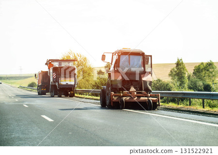 A tractor with a brush sweeps the road against the backdrop of a milling machine cutting asphalt. Country road repairs. Copy space for text, industry A tractor with a brush sweeps the road against the backdrop of a milling machine cutting asphalt. Country road repairs. Copy space for text, industry 131522921
