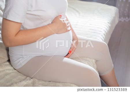 Pregnant woman relaxing at home in comfortable clothing while cradling her baby bump Pregnant woman relaxing at home in comfortable clothing while cradling her baby bump 131523389