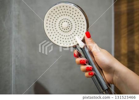 female hand with red manicure holding shower head in hand against grey tile background in bathroom, close-up female hand with red manicure holding shower head in hand against grey tile background in bathroom, close-up 131523466