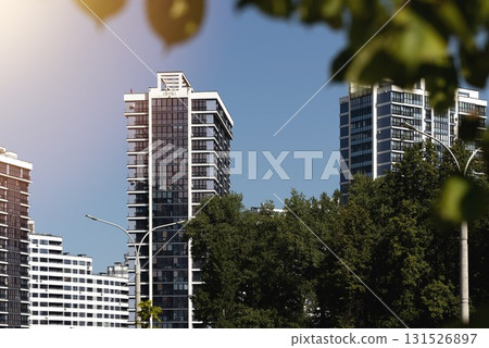 Sunny panoramic view of modern residential tower with glass facade surrounded by trees and greenery. 131526897