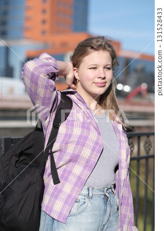 young woman smiles softly, holding a backpack while standing near a modern urban building. The photo conveys a blend of casual confidence and city life exploration. young woman smiles softly, holding a backpack while standing near a modern urban building. The photo conveys a blend of casual confidence and city life exploration. 131528433