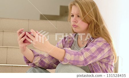 A blonde girl in a plaid shirt furrows her brow while using her smartphone, reflecting the intensity of digital interactions. The scene underscores the challenges of modern connectivity. 131528435