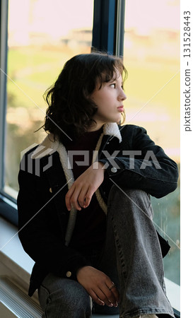 A contemplative girl sits by a window, her gaze directed outward. The photo highlights a reflective mood, symbolizing personal growth and the beauty of stillness. 131528443