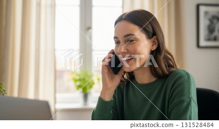 A smiling woman talking on the phone while working from home with a laptop. Cheerful professional having a pleasant conversation in a home office 131528484