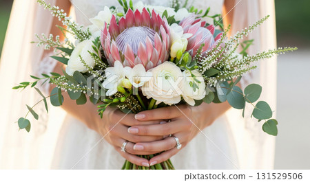 A hyper-realistic, high-detail close-up photograph of a bride's hands gently holding a gorgeous wedding bouquet filled with pale pink protea 'Blushing Bride', ivory Freesias, soft foliage 131529506
