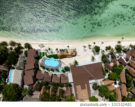 Drone view of a resort featuring a pool, palm trees, and a turquoise beach under a sunny sky. Rin Nai Beach. Ko Pha Ngan, Thailand. Drone view of a resort featuring a pool, palm trees, and a turquoise beach under a sunny sky. Rin Nai Beach. Ko Pha Ngan, Thailand. 131532481