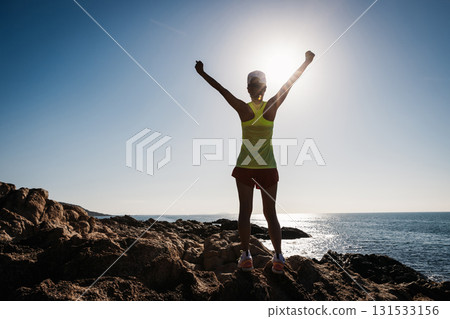 Athletic woman open arms to sunrise on seaside rocks 131533156
