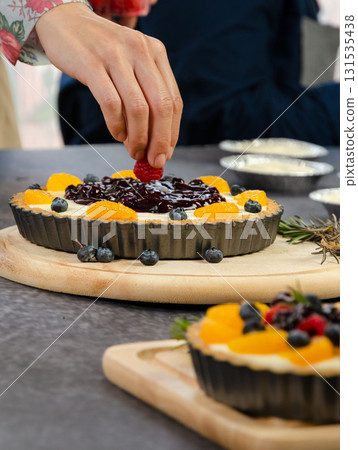 Baker carefully place fresh raspberry on fruit cheese pie of orange and blueberries arranged on creamy filling in fluted pastry tray Baker carefully place fresh raspberry on fruit cheese pie of orange and blueberries arranged on creamy filling in fluted pastry tray 131535438