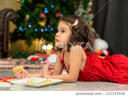 Young girl in red dress lies on floor near decorated Christmas tree while holding festive cookie in living room at home 131535444
