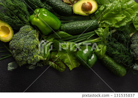 Various green vegetables such as broccoli, cucumbers, green peppers, and lettuce, fresh herbs like basil and dill, along with ripe avocado are artfully arranged on a dark background, healthy meal Various green vegetables such as broccoli, cucumbers, green peppers, and lettuce, fresh herbs like basil and dill, along with ripe avocado are artfully arranged on a dark background, healthy meal 131535837