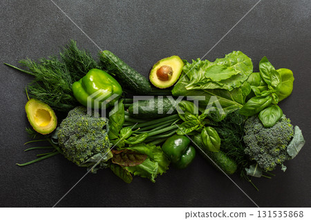 A vibrant assortment of fresh green vegetables is carefully arranged on a dark surface This selection includes broccoli, green bell peppers, cucumbers, spinach, lettuce, and an avocado, healthy eating 131535868