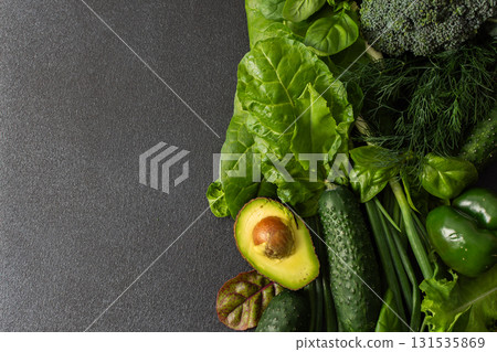 Displaying a vibrant assortment of fresh greens and vegetables on a dark surface, this scene features cucumbers, leafy lettuce, broccoli, green peppers, and an avocado cut in half, a healthy lifestyle 131535869