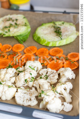 Cauliflower, carrots, and cabbage are sliced and arranged on a tray lined with parchment paper, ready for roasting with herbs in a warm kitchen setting 131535946