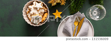 A festive table setting with an empty plate, napkin and cutlery and gingerbread on a green background. Spruse branches and golden tree ornaments enhance the holiday ambiance, banner A festive table setting with an empty plate, napkin and cutlery and gingerbread on a green background. Spruse branches and golden tree ornaments enhance the holiday ambiance, banner 131536009