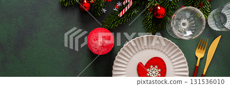 A cozy Christmas table with an empty plate, cutlery and candle on a rich green background. Red Christmas tree decorations and spruse branches complete the festive look, banner 131536010