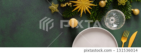 A Christmas table setting with an empty plate and cutlery on a green background. Spruse branches and golden tree decorations add elegance and festive cheer to the scene, banner 131536014