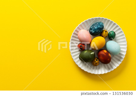 A variety of beautifully painted Easter eggs in different colors and patterns are placed on a delicate white plate. The vibrant hues stand out against the cheerful yellow backdrop 131536030