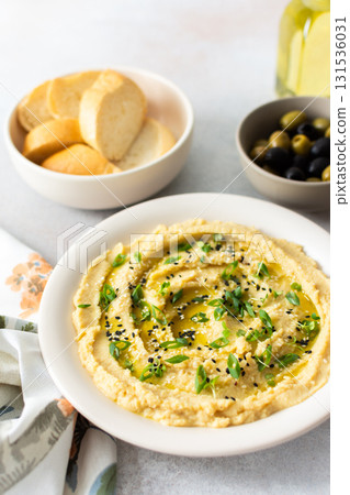 A bowl of creamy hummus topped with olive oil and herbs sits beside fresh bread and a bowl of olives. The light setting showcases a simple, inviting meal A bowl of creamy hummus topped with olive oil and herbs sits beside fresh bread and a bowl of olives. The light setting showcases a simple, inviting meal 131536031