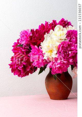 A vibrant arrangement of pink and white peonies fills a ceramic vase, placed on a soft pink tablecloth. The flowers add a cheerful touch to any room, ideal for spring decor 131536037