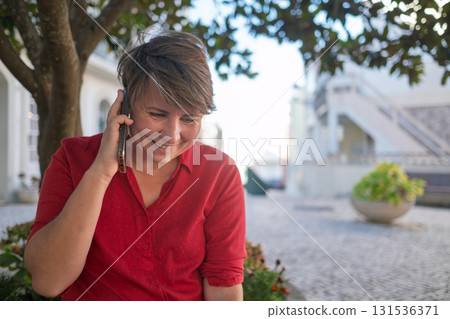 European woman sitting outdoors in the shade, talking on the phone and laughing 131536371