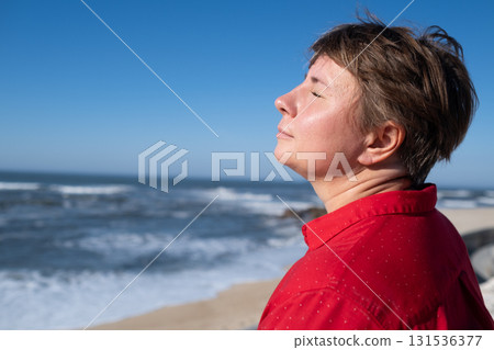 Middle-aged woman sitting on a wall looking at the ocean in Portugal, enjoying a sunny summer day 131536377