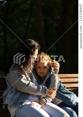Candid portrait of happy mother and son sitting on bench in public park on sunny summer day. Holidays with kids. Parenthood and motherhood topics. Candid portrait of happy mother and son sitting on bench in public park on sunny summer day. Holidays with kids. Parenthood and motherhood topics. 131536497