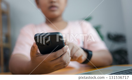 Close up of child using smartphone with smartwatch and laptop on table, digital lifestyle concept Close up of child using smartphone with smartwatch and laptop on table, digital lifestyle concept 131536717