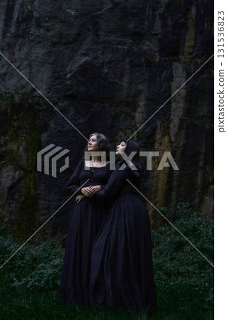 two women in the black long dresses sitting on black rocks on Halloween, performing a witch ritual, modern witch two women in the black long dresses sitting on black rocks on Halloween, performing a witch ritual, modern witch 131536823