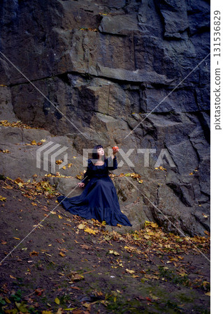 woman in a black long dress sits on black rocks on Halloween holding a pumpkin-shaped cup, a modern witch 131536829