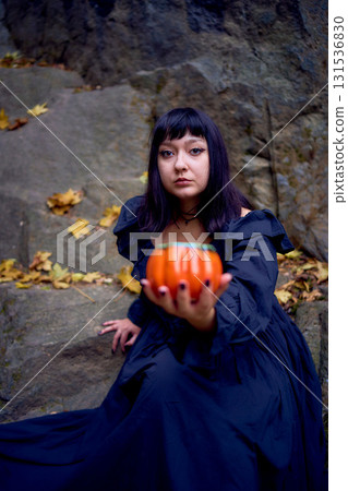 woman in a black long dress sits on black rocks on Halloween holding a pumpkin-shaped cup, a modern witch 131536830