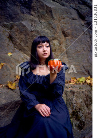 woman in a black long dress sits on black rocks on Halloween holding a pumpkin-shaped cup, a modern witch woman in a black long dress sits on black rocks on Halloween holding a pumpkin-shaped cup, a modern witch 131536831