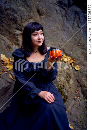 woman in a black long dress sits on black rocks on Halloween holding a pumpkin-shaped cup, a modern witch woman in a black long dress sits on black rocks on Halloween holding a pumpkin-shaped cup, a modern witch 131536832